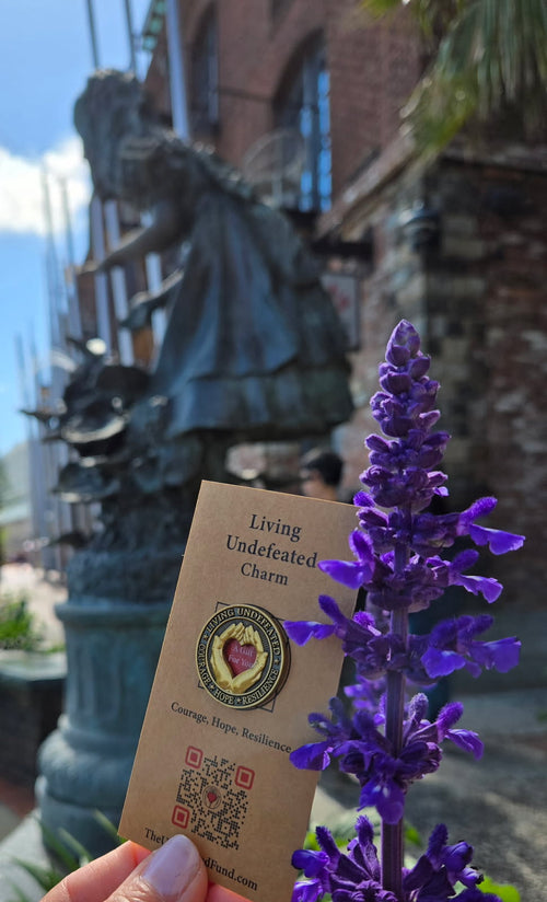 A Living Undefeated Charm held up beside purple flowers and a bronze statue