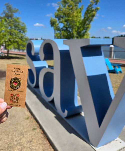 A Living Undefeated Charm held in front of a large blue letter sculpture on a waterfront