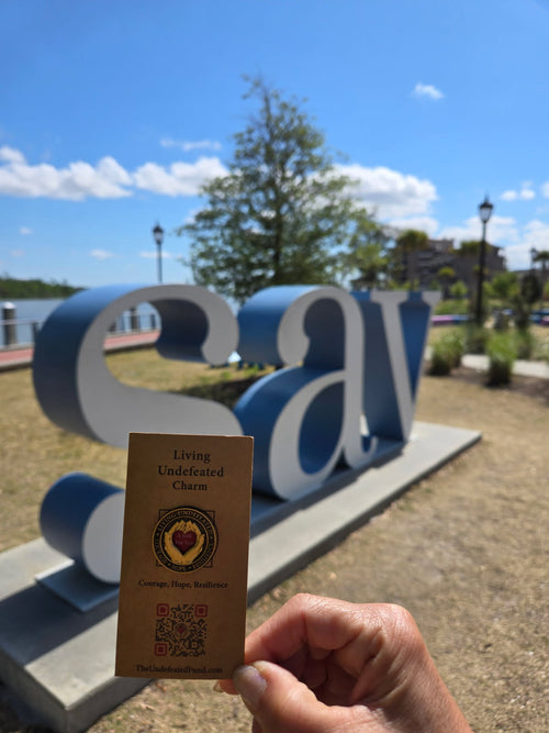 A Living Undefeated Charm held beside a riverfront letter sculpture under a blue sky